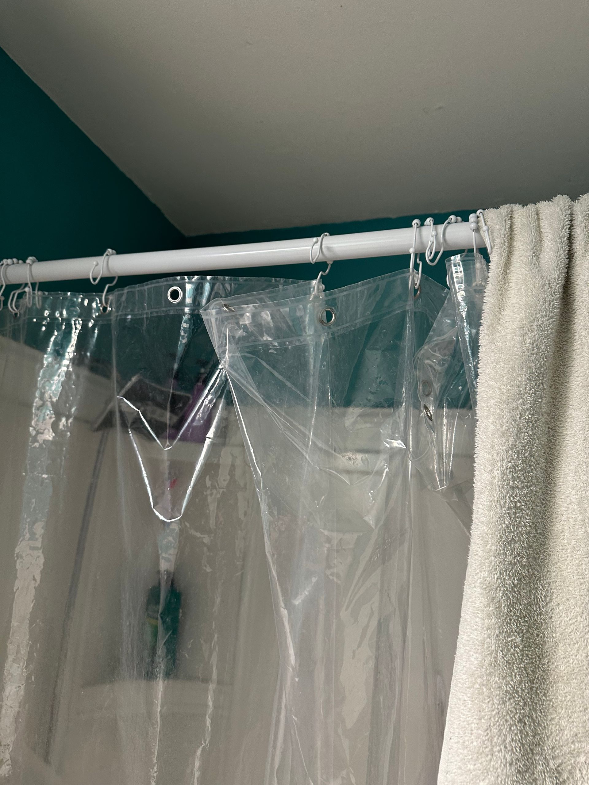 Clear shower curtain hanging from a white rod beside a beige curtain, under a sloped ceiling.