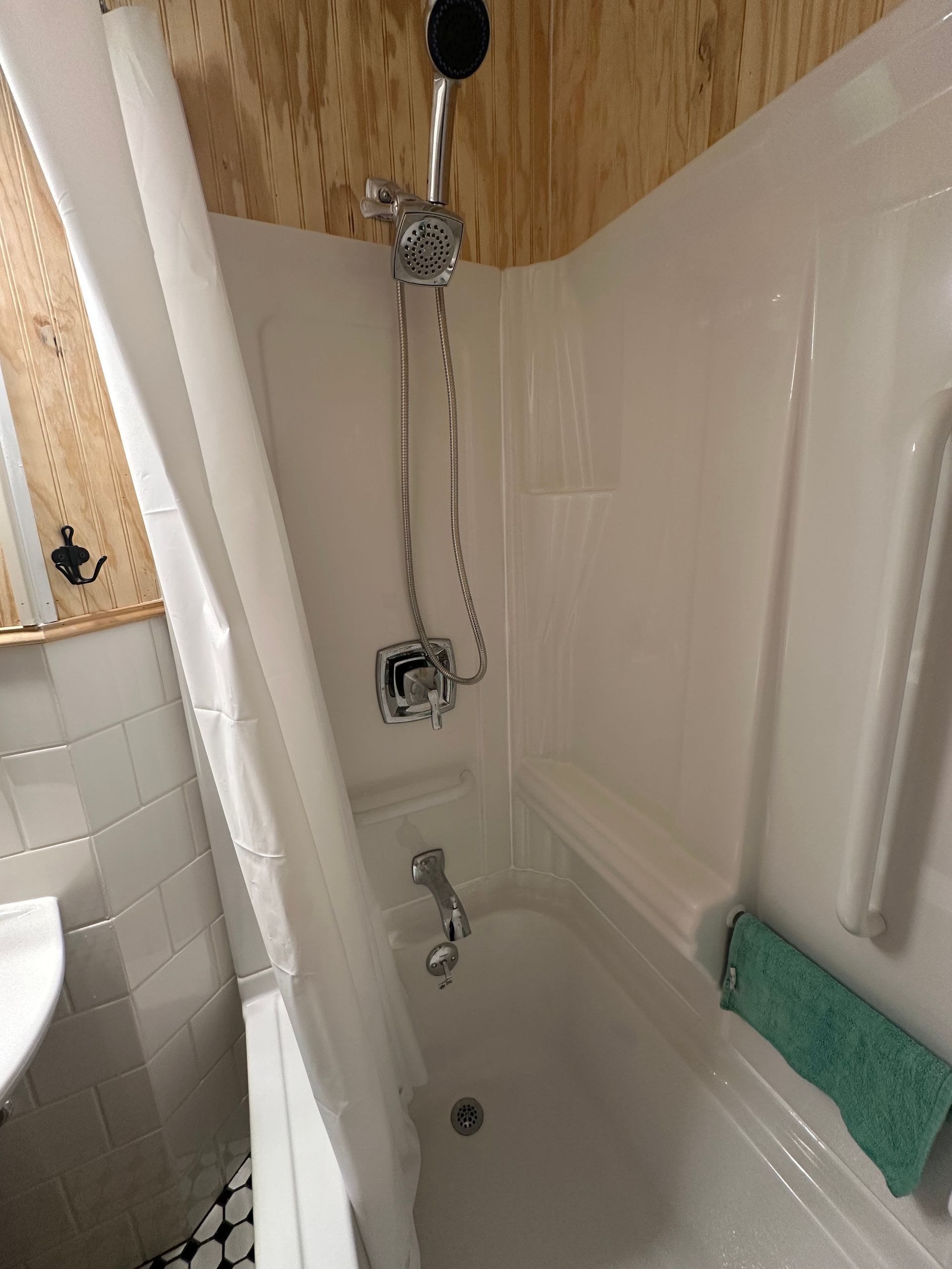 Small bathroom shower stall with tub, handheld shower, white tile, and green towel