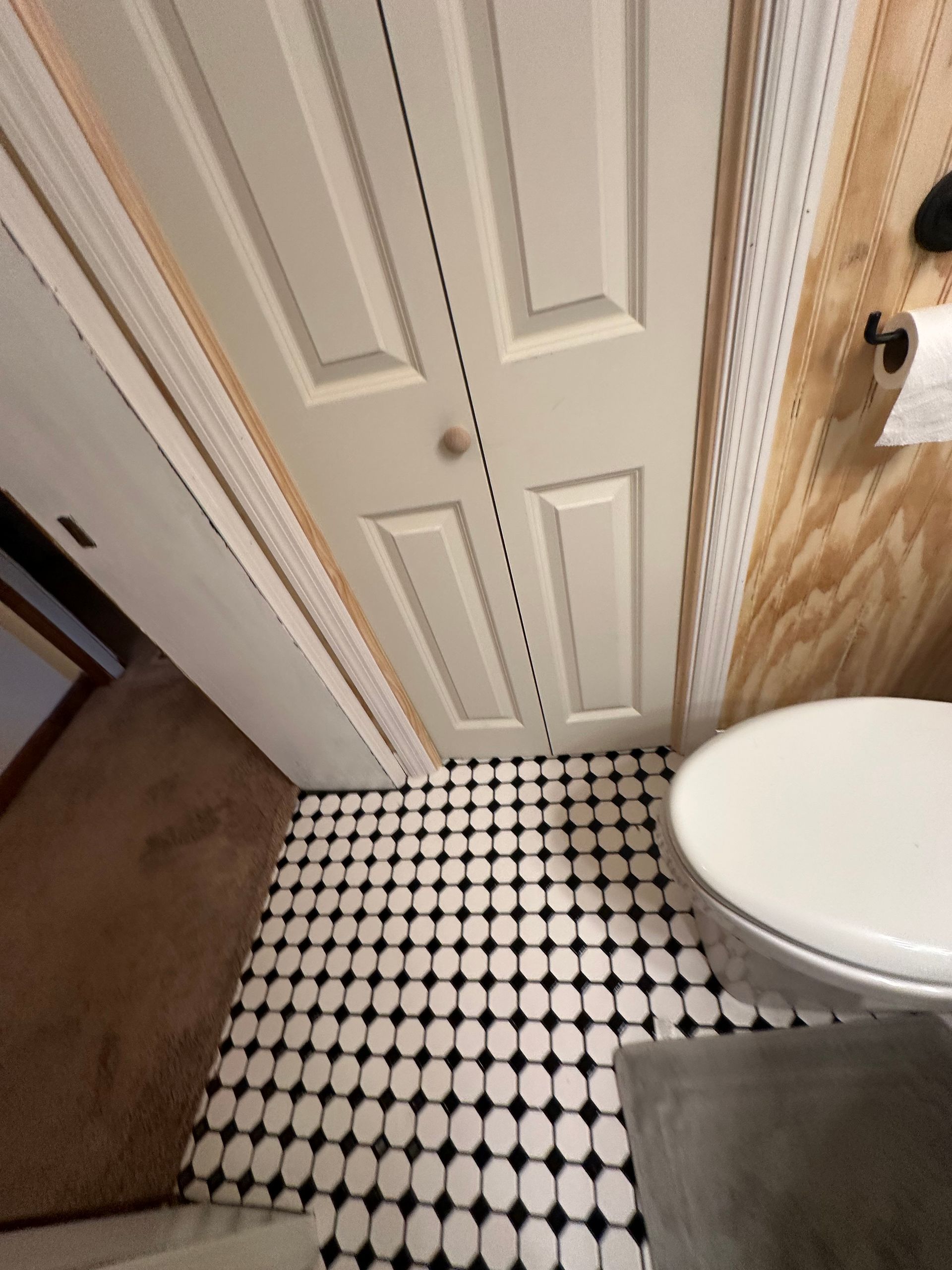 Narrow bathroom corner with white paneled door, black-and-white tile floor, toilet, and shower edge