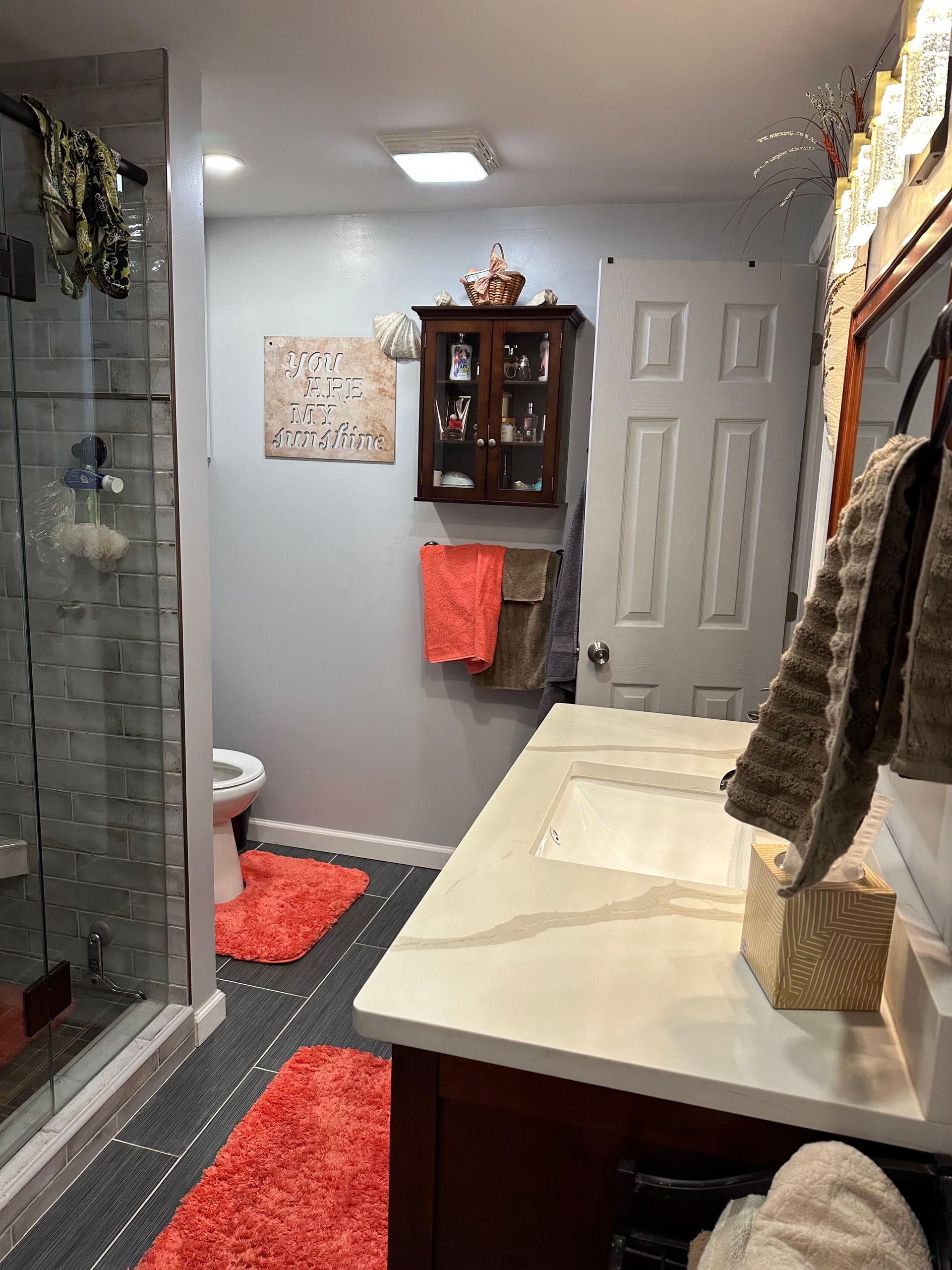 Bathroom with glass shower, vanity, mirror, and red rugs on a gray floor.