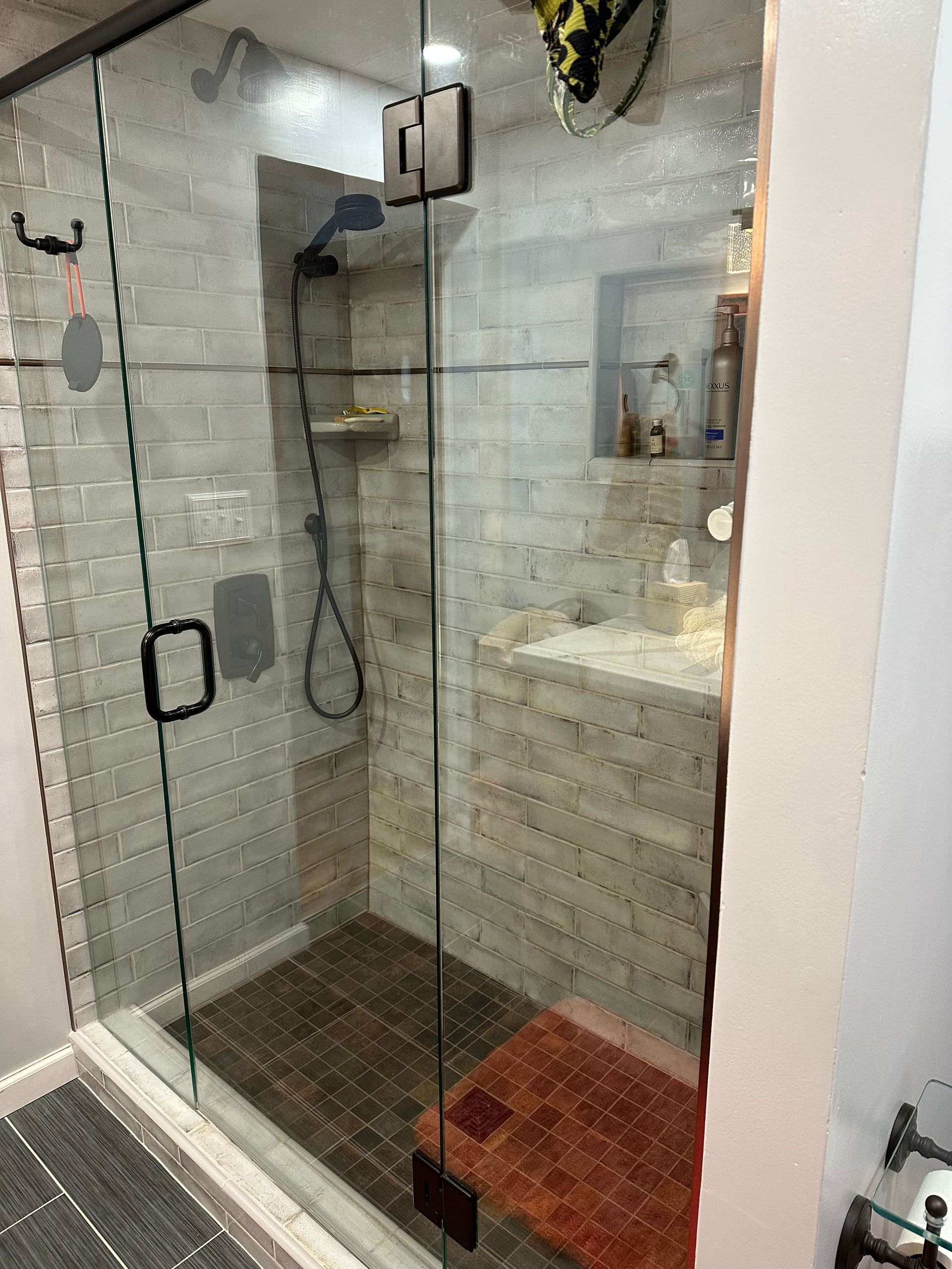 Glass shower stall with rainfall showerhead, gray tile walls, and a red bath mat outside the door