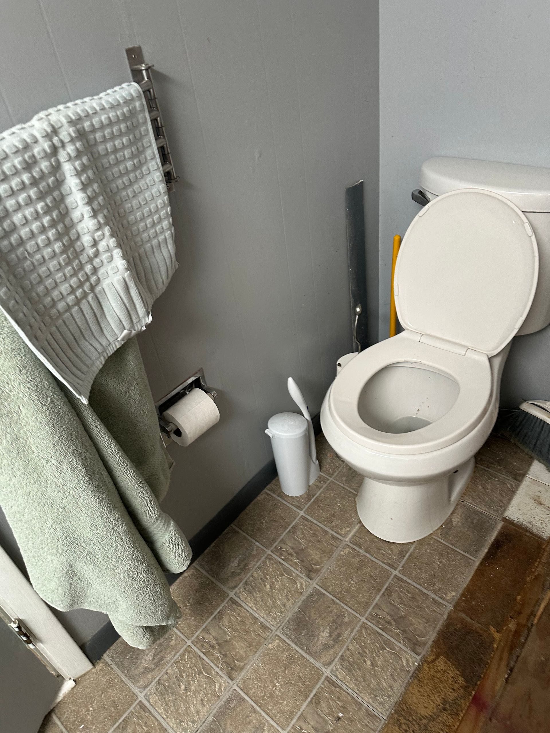 Small bathroom with an open toilet, gray towels, white cup, and tiled floor.
