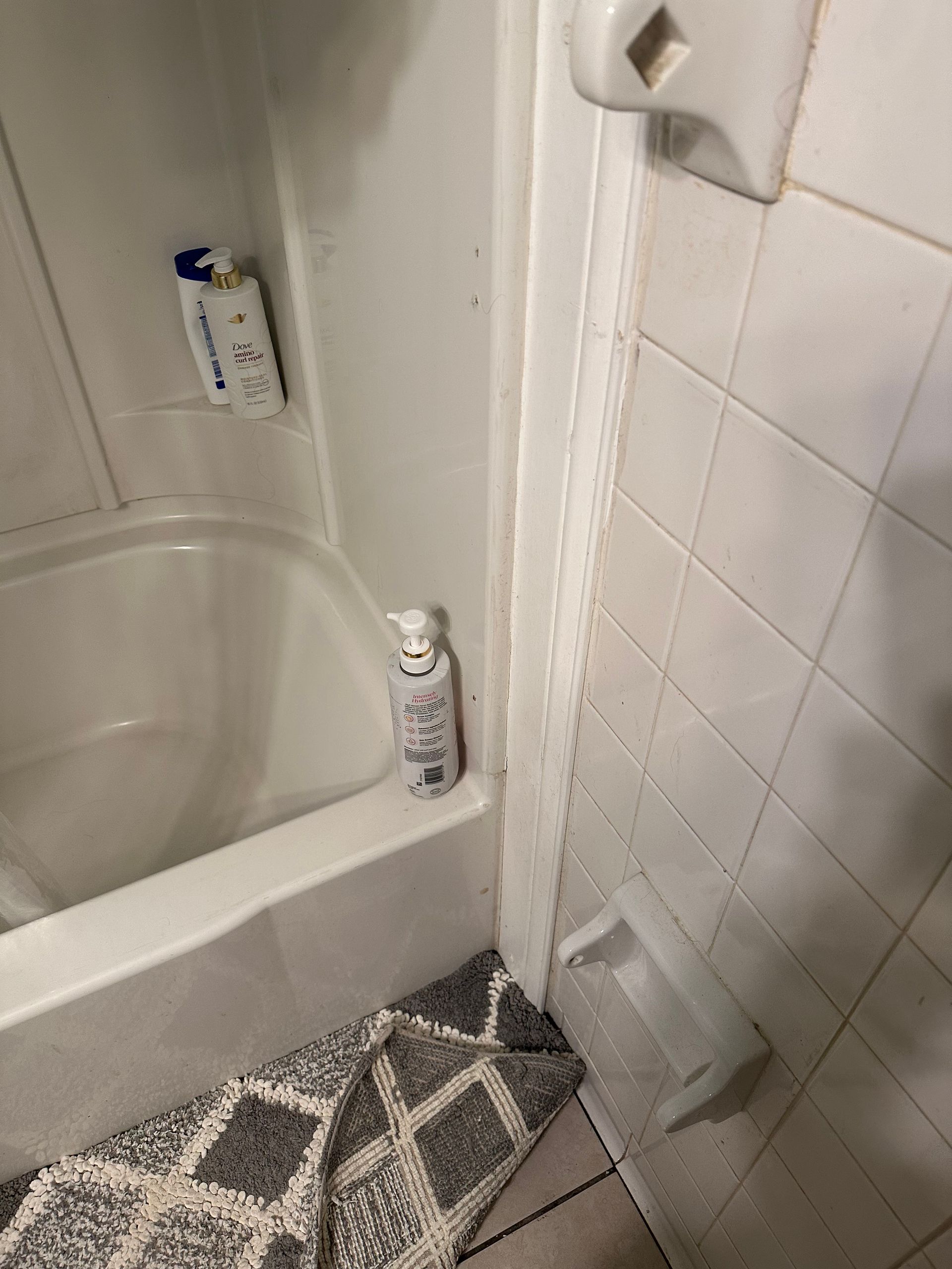 Small bathroom corner with a bathtub, white tiled wall, rug, and two bottles on the tub ledge and floor.