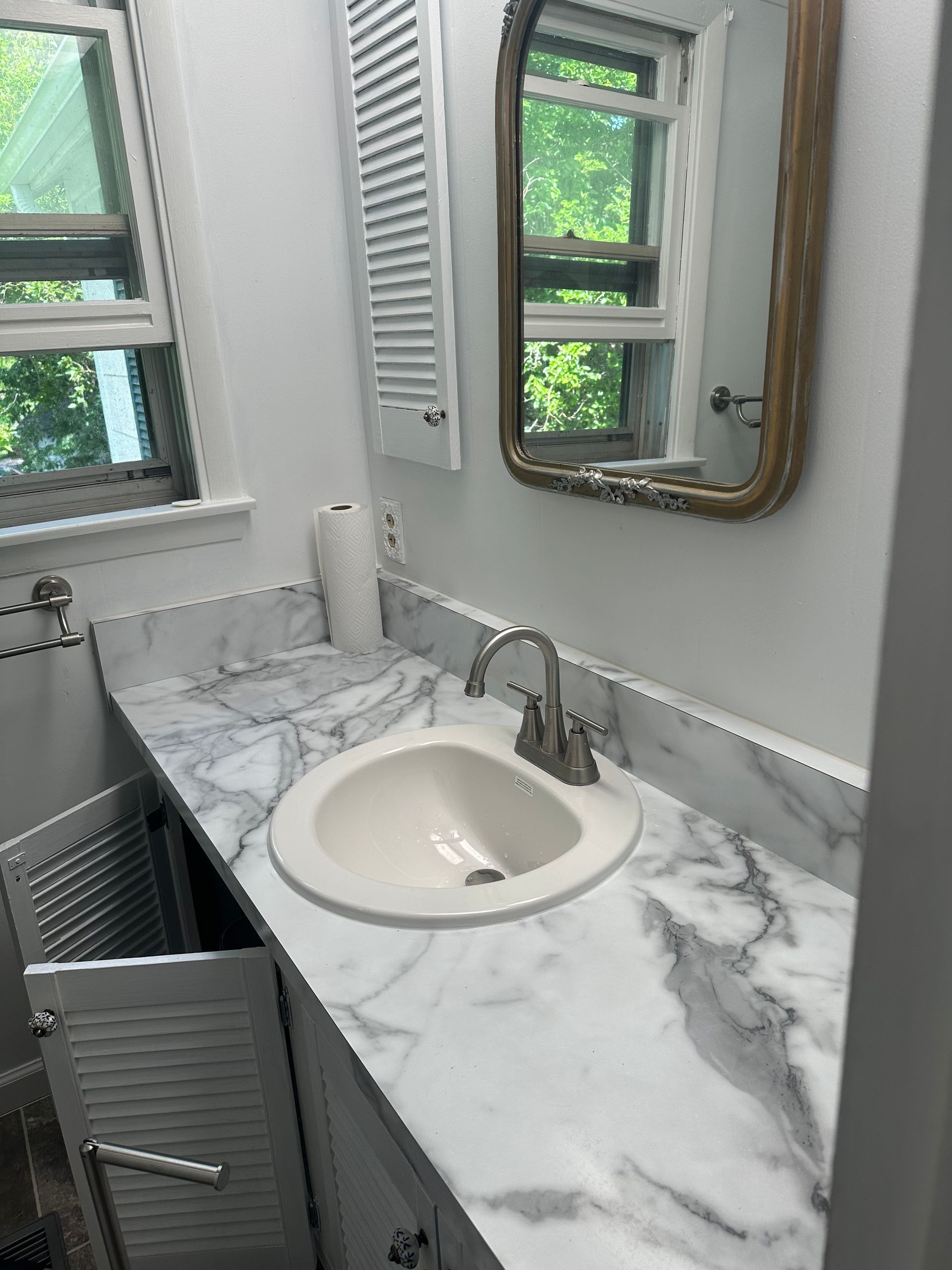 Small bathroom vanity with marble countertop, oval sink, gray cabinets, mirror, and windows.