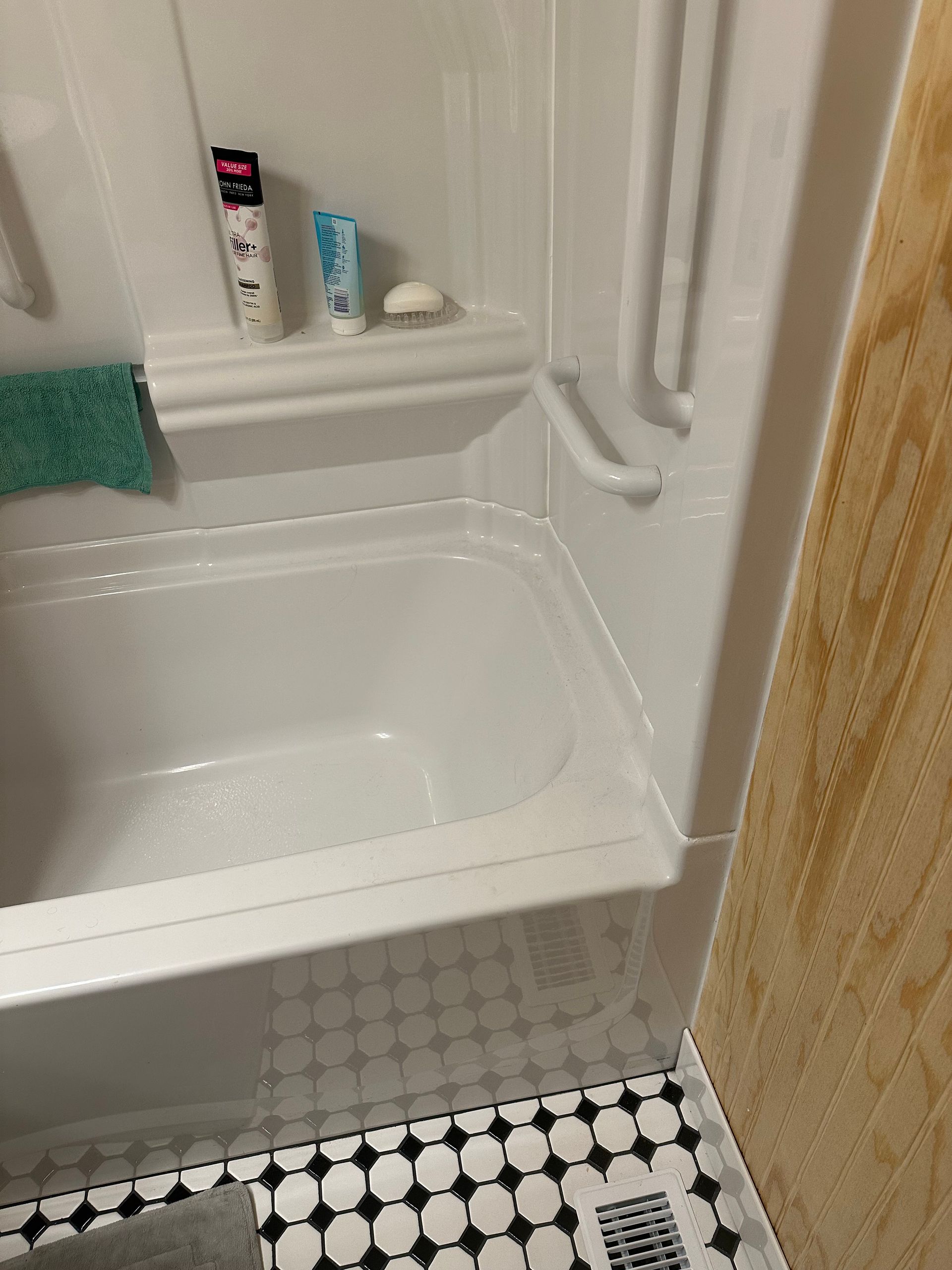 Small bathroom with a white bathtub, teal towel, shower items, and black-and-white tiled floor.