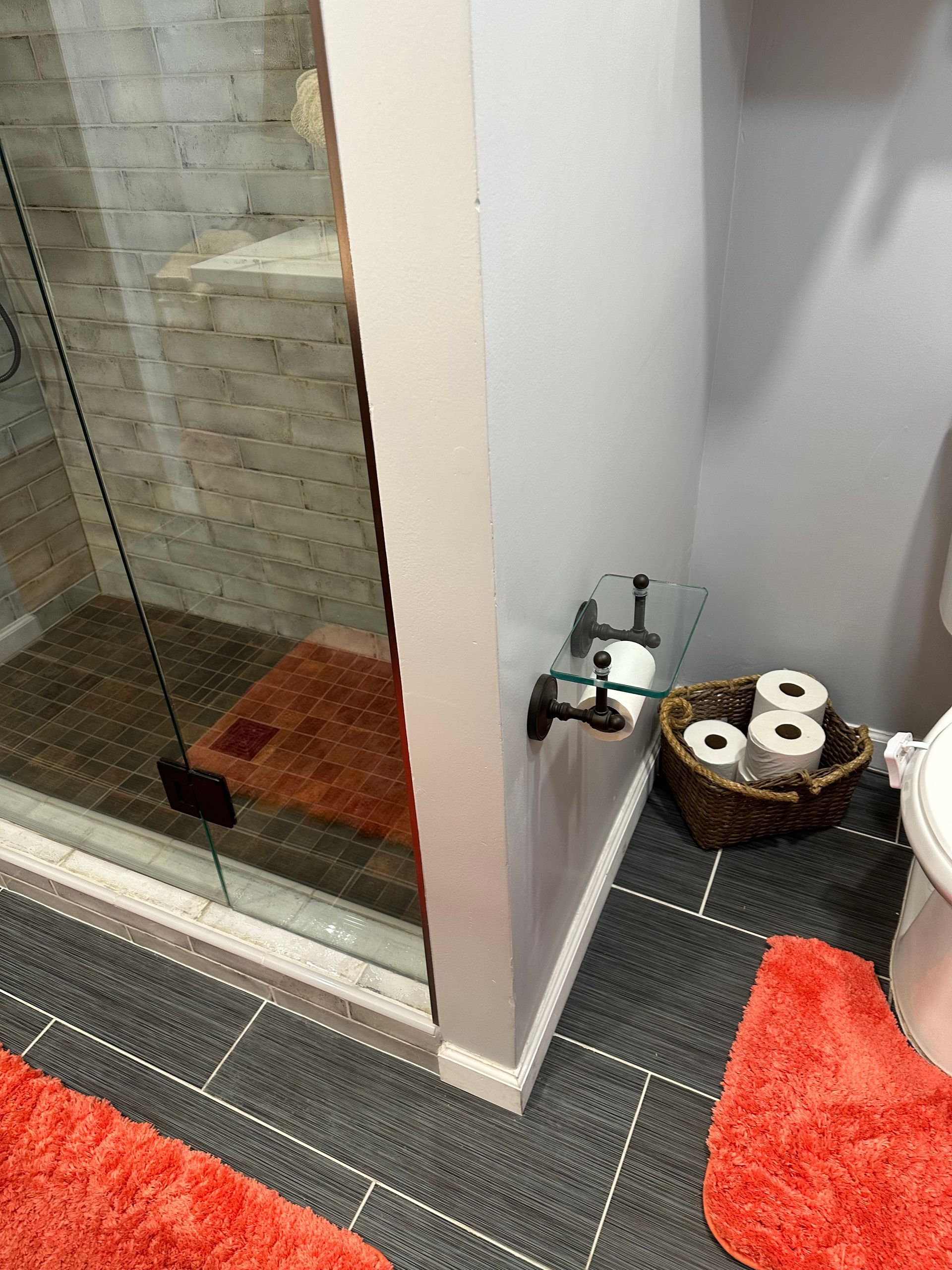 Bathroom corner with glass shower, dark tile floor, toilet paper basket, and red bath mat