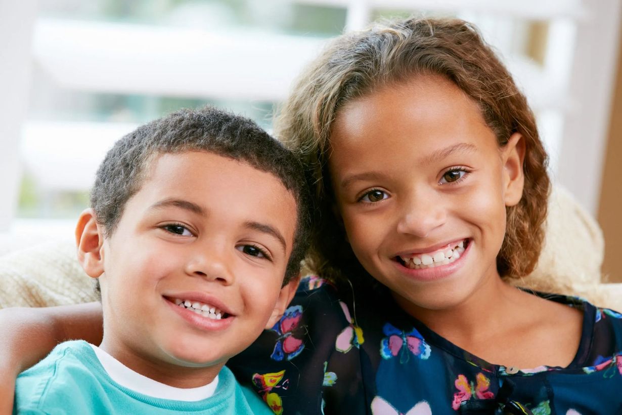 Two smiling children sitting close together indoors, wearing casual clothing.
