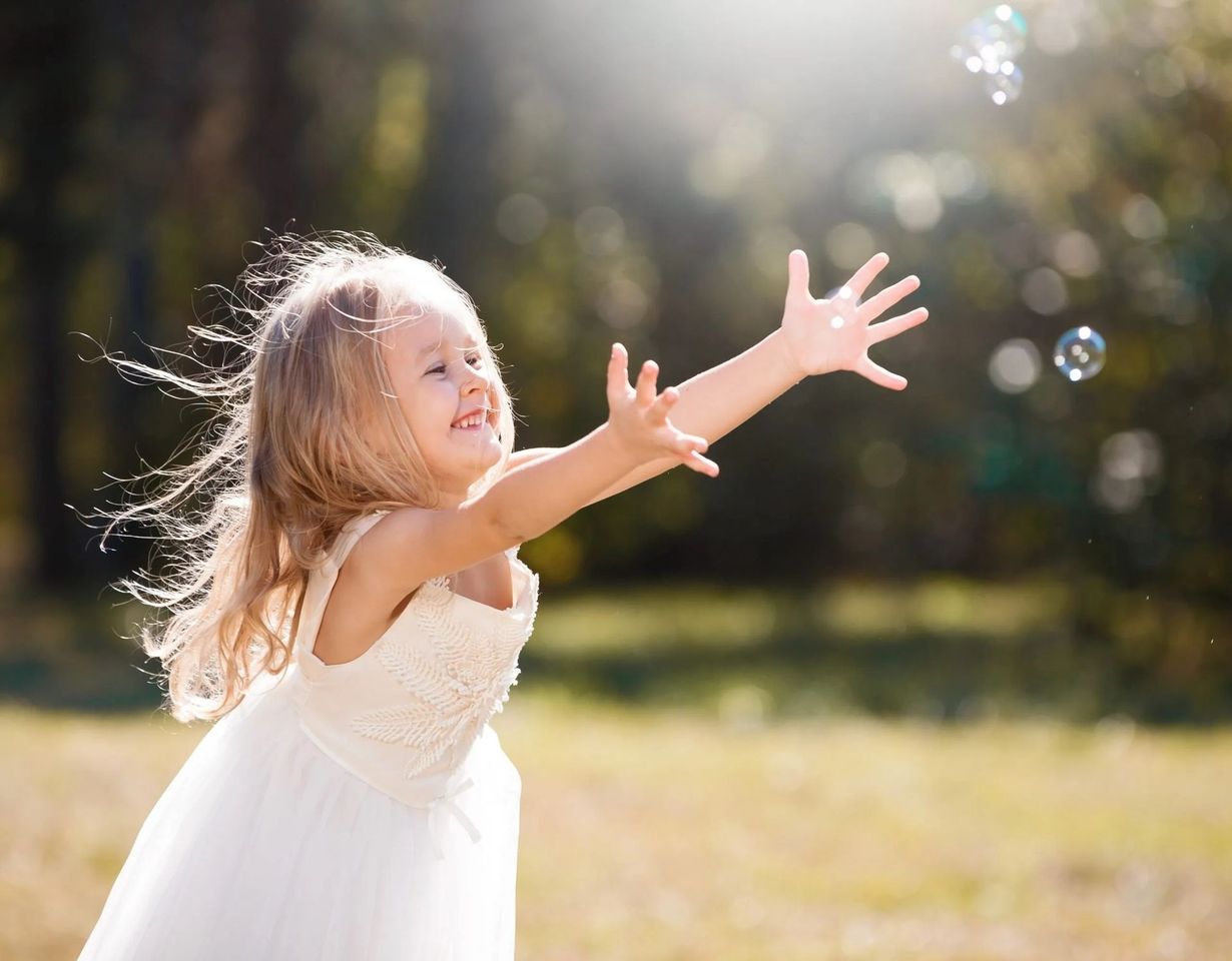 A young child in a white dress reaches toward floating bubbles in a sunlit, grassy outdoor setting.