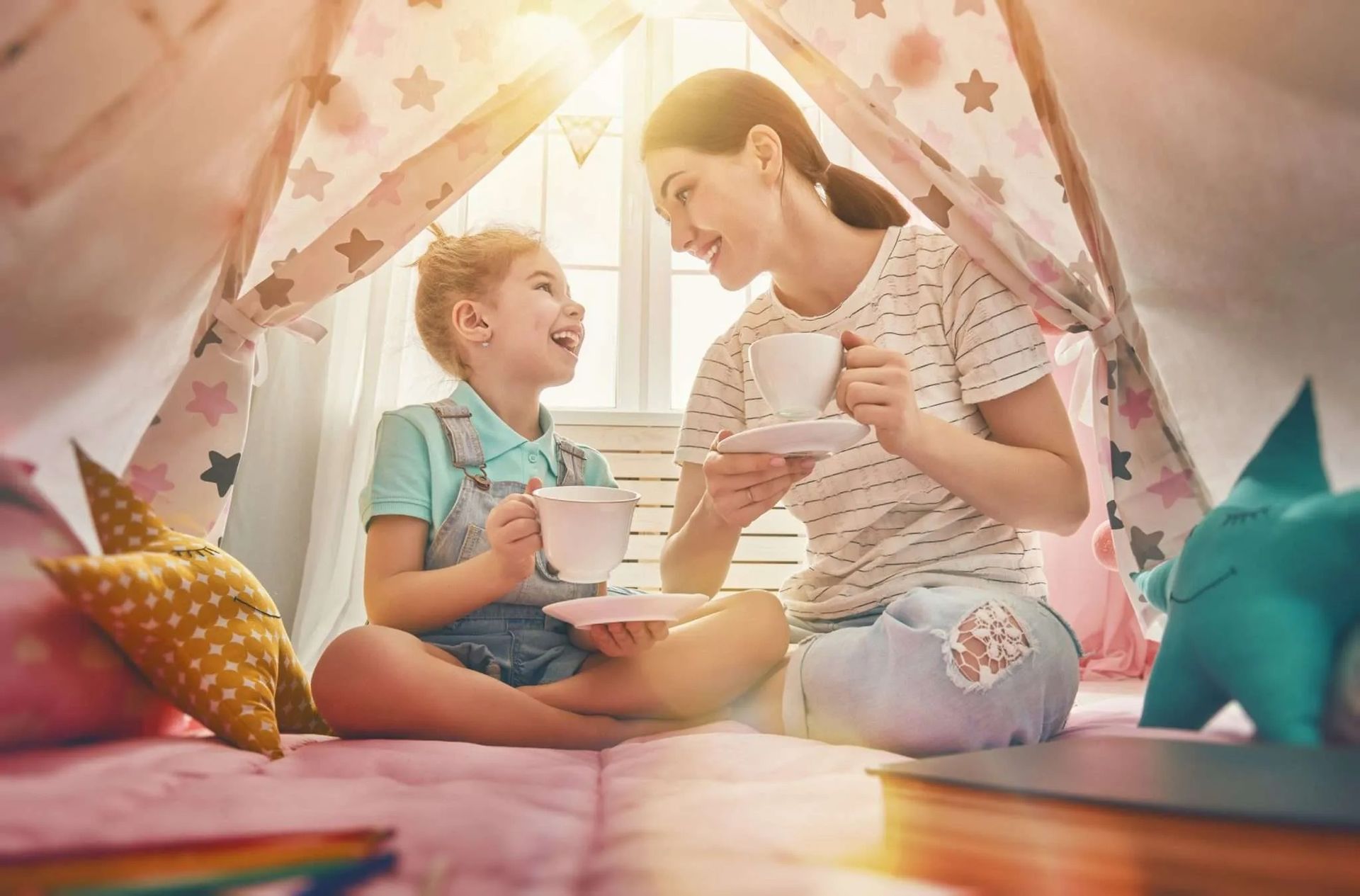 A child and an adult sitting inside a star-patterned tent, smiling while holding toy teacups in a warm, sunlit room.