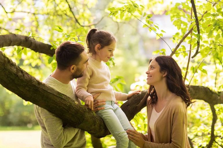 A family of three interacts lovingly while sitting among the branches of a tree in a sunny, green outdoor setting.