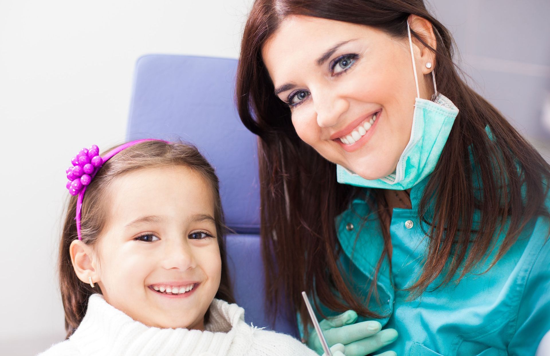 A smiling dentist in scrubs and a mask poses next to a young patient in a dental chair.