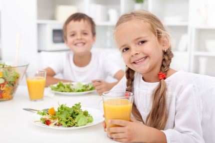 Two children smile at a table with plates of salad and glasses of orange juice in a bright, white kitchen.