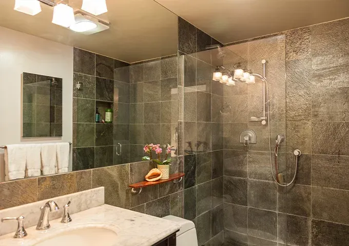 Modern bathroom with marble sink, glass shower, gray tile walls, and vanity mirror lighting
