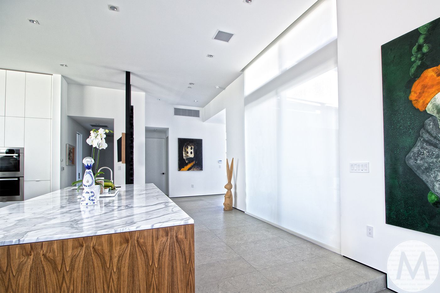 Modern kitchen with marble island, wood base, white walls, art, and sheer window coverings.