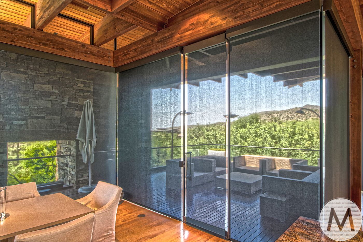 Patio with dark blinds, outdoor furniture, stone fireplace, and wooden ceiling, with view of greenery.