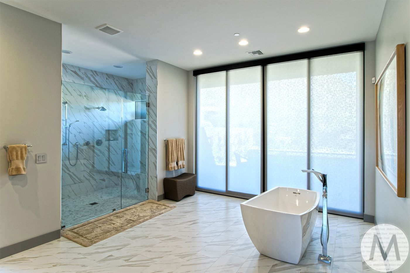 Modern bathroom with marble floors, glass shower, soaking tub, and large window shades.
