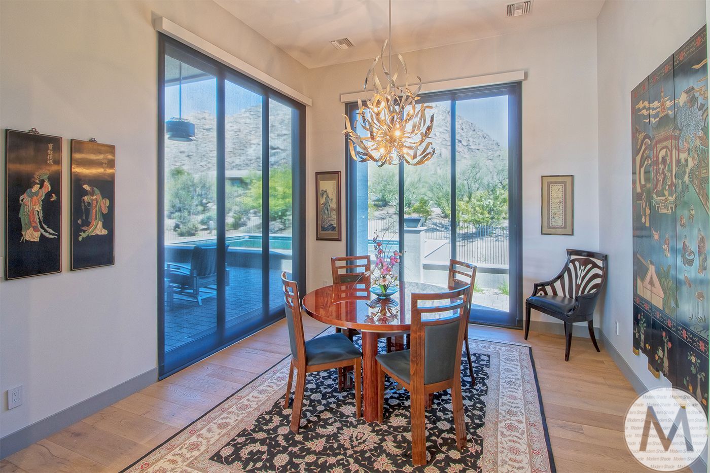 Dining room with round table, chairs, chandelier, and large windows overlooking a landscape.