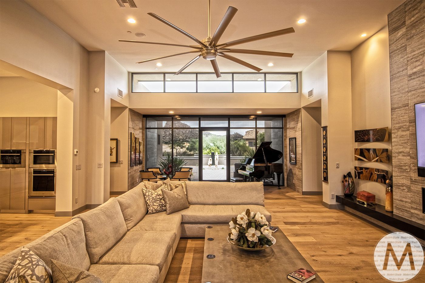 Large, open living room with wood floors, a sectional sofa, a grand piano, and a large ceiling fan.