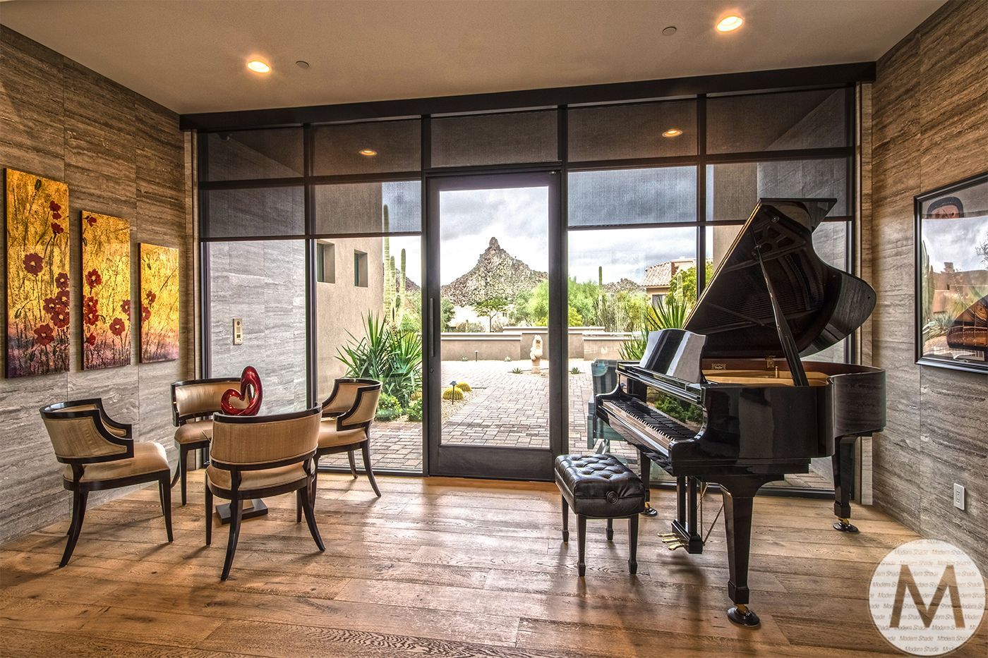 A room with a grand piano, small table with chairs, and large windows overlooking a desert landscape.