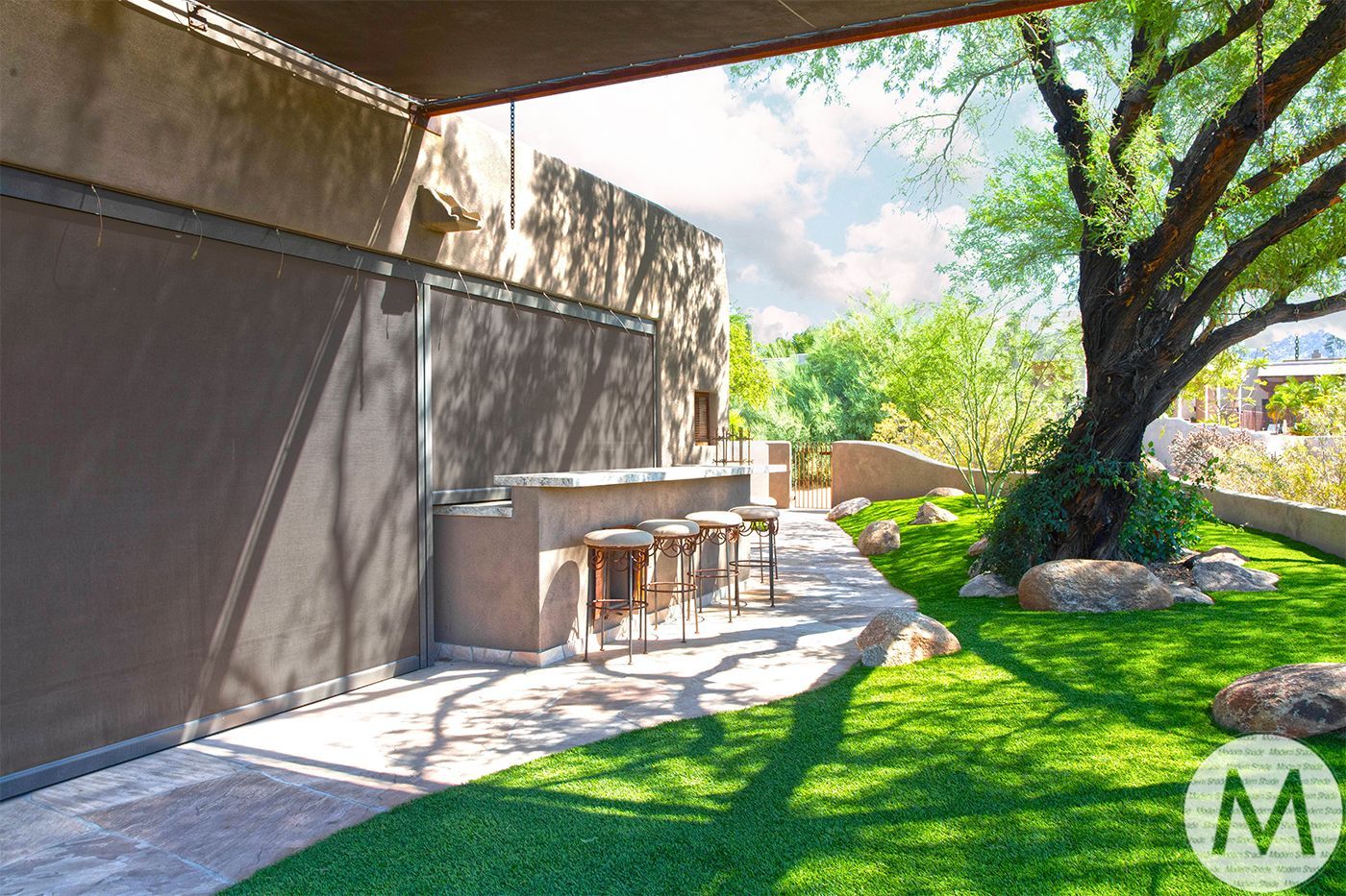 Outdoor bar with stools on patio, lush green lawn, and large tree with sunlight.