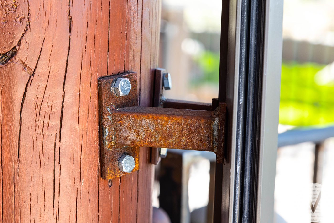 Rusty metal latch on a wooden post, bolted to a window frame, outdoors.