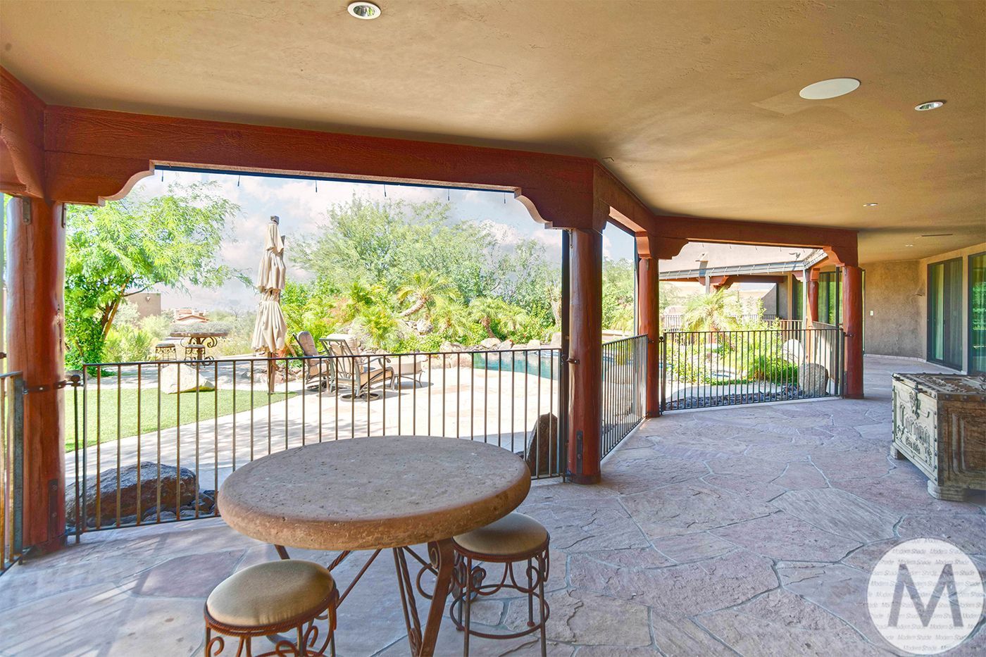 Covered patio with stone table and wrought-iron railing overlooking backyard with pool.