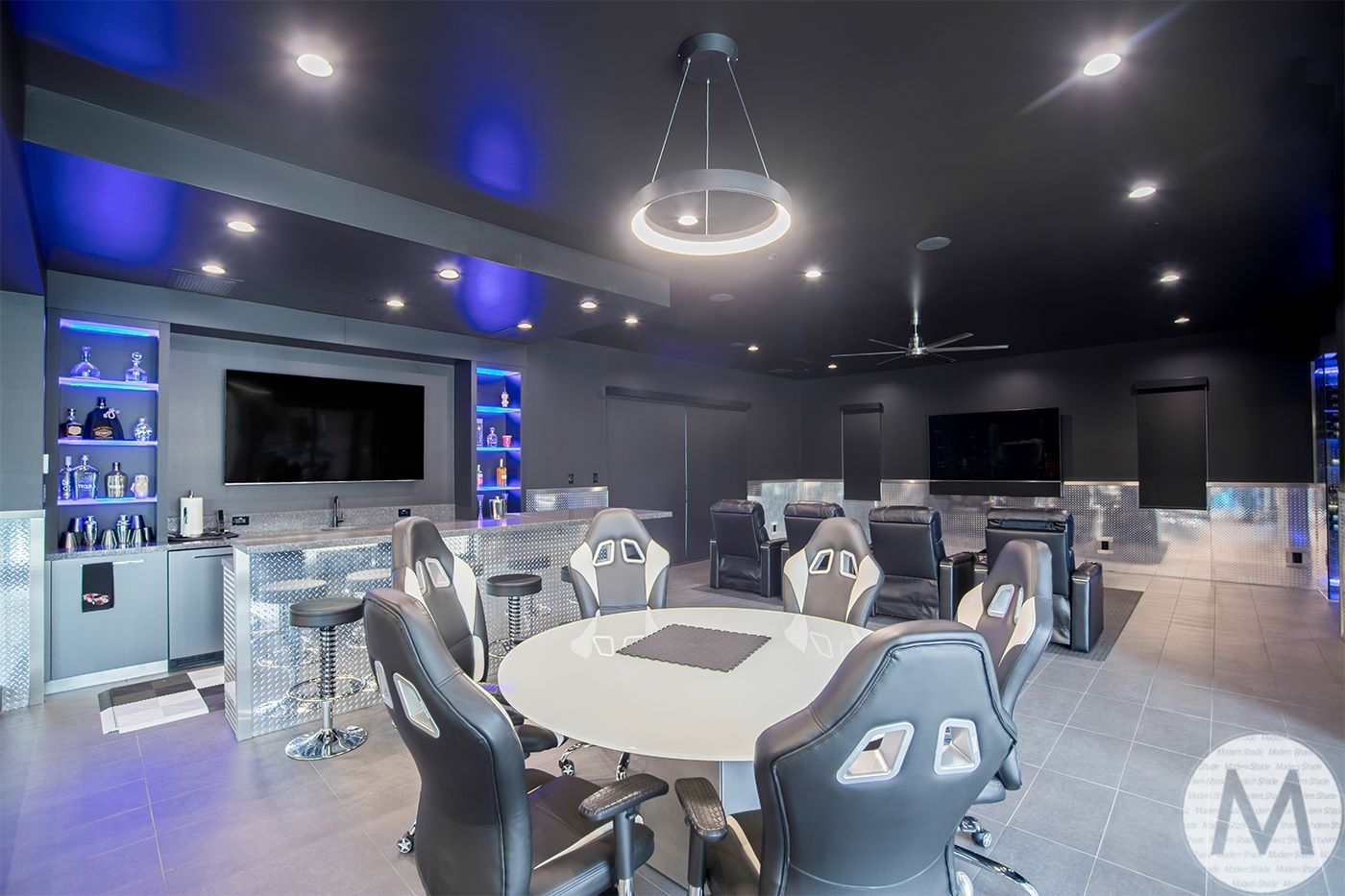 Gaming room with gaming chairs around a table, bar, and large screens.