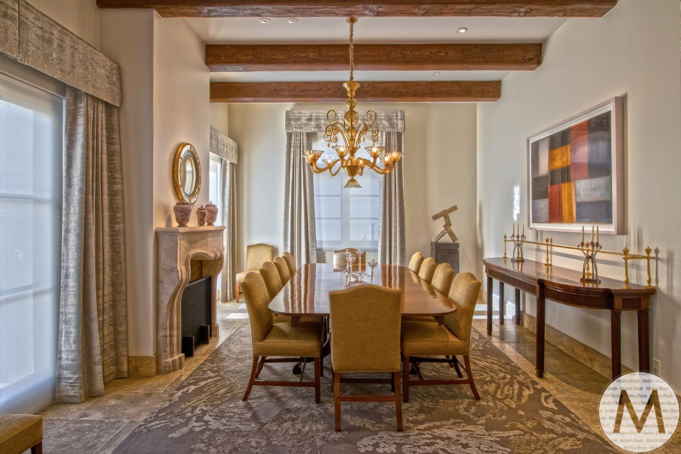 Elegant dining room with long table, upholstered chairs, fireplace, artwork, and chandelier.