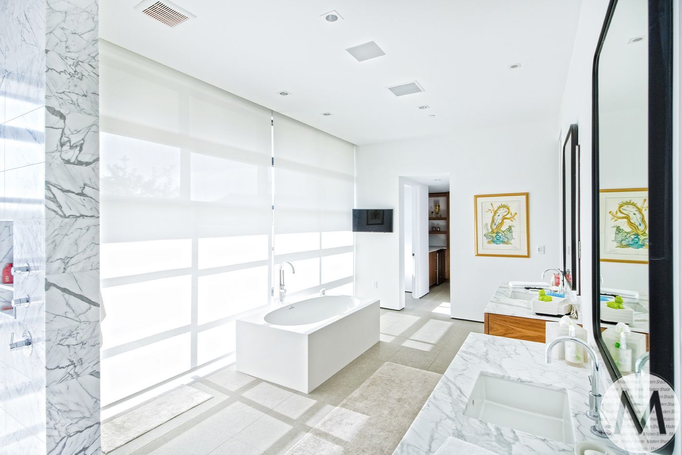 Bright white bathroom with a tub by a large window, marble countertop, and a television.