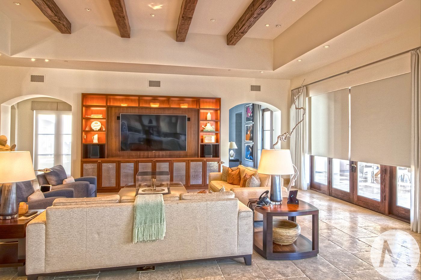 Living room with TV built-in to wooden wall, arched doorways, large windows, wood beams, and neutral colors.