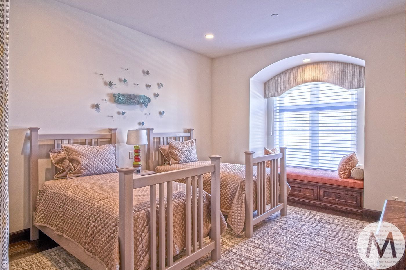 Two twin beds in a light-toned bedroom with window seat, wall decor, and a patterned rug.