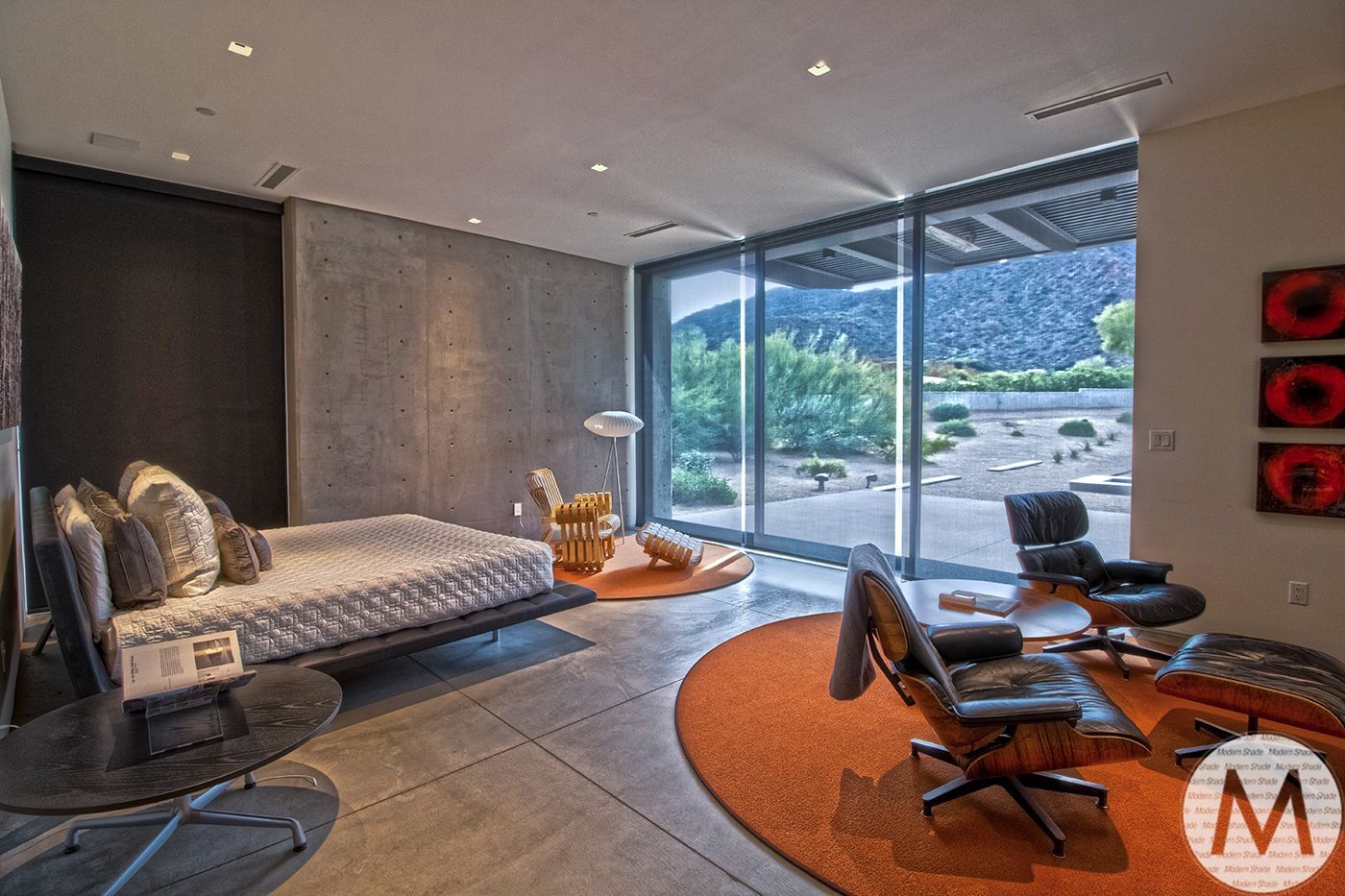 Modern bedroom with bed, seating, large window, concrete walls, and an orange rug.