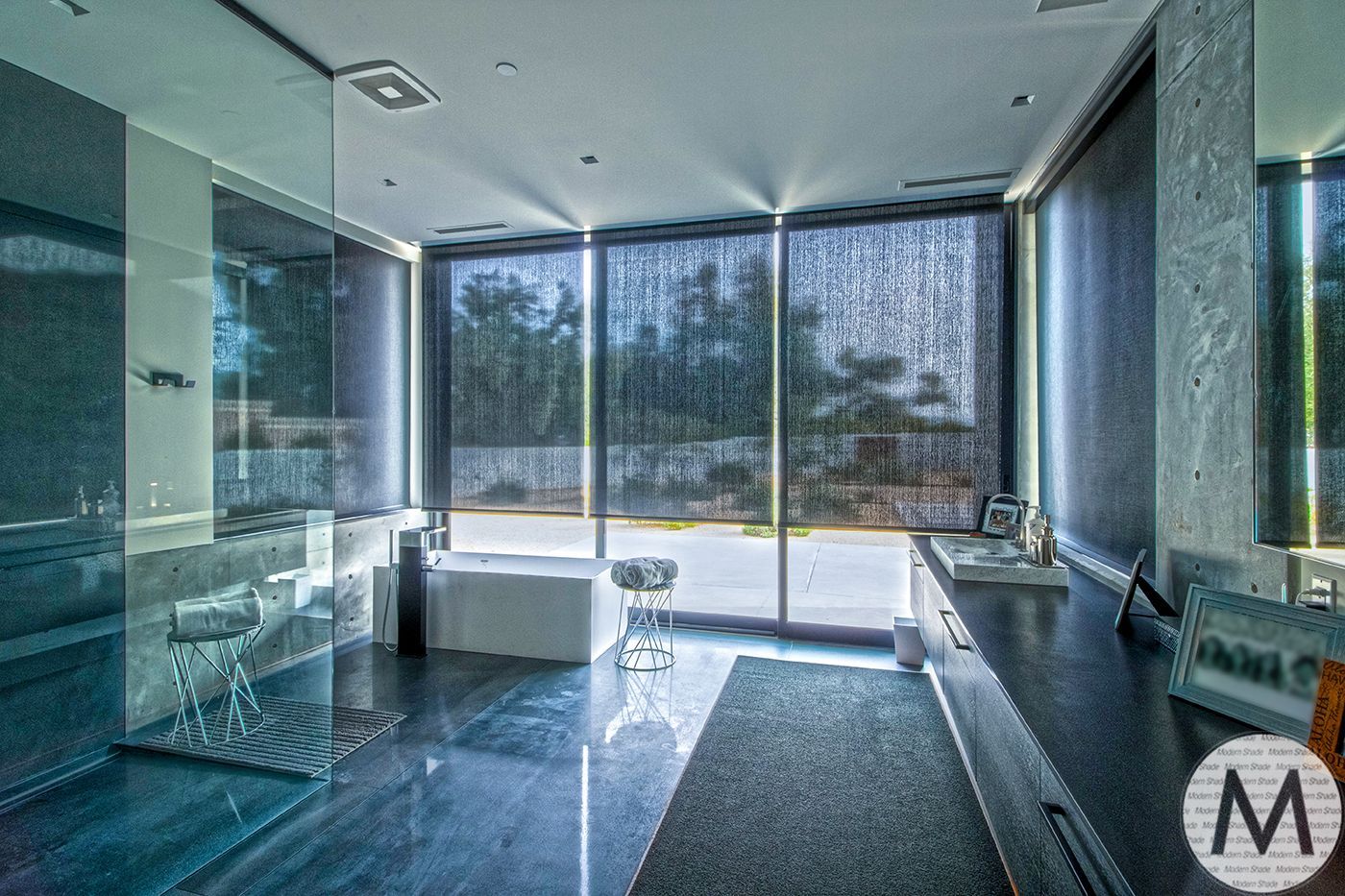 Modern, dark-toned bathroom with large windows, a bathtub, and a long countertop.