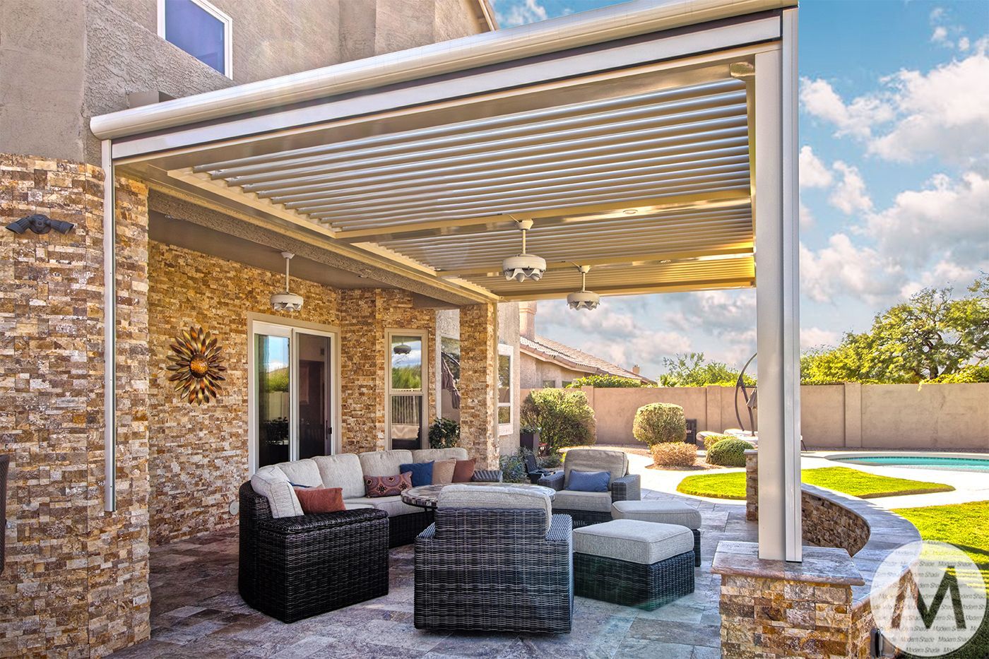 Patio with stone walls, furniture, and adjustable louvered roof overlooking a pool and yard.