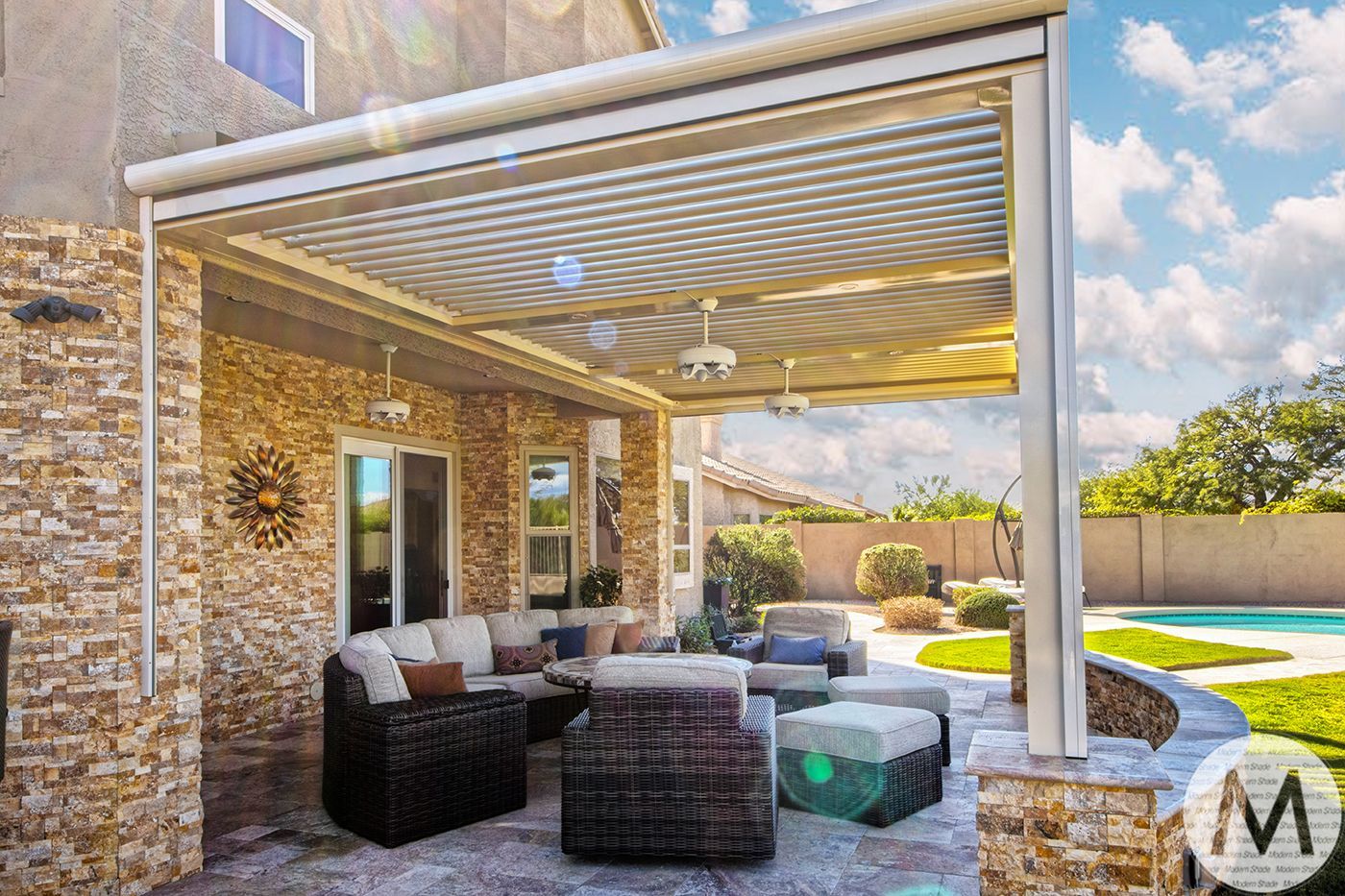 Patio with beige stone wall and outdoor furniture under a louvered awning, pool in the background.