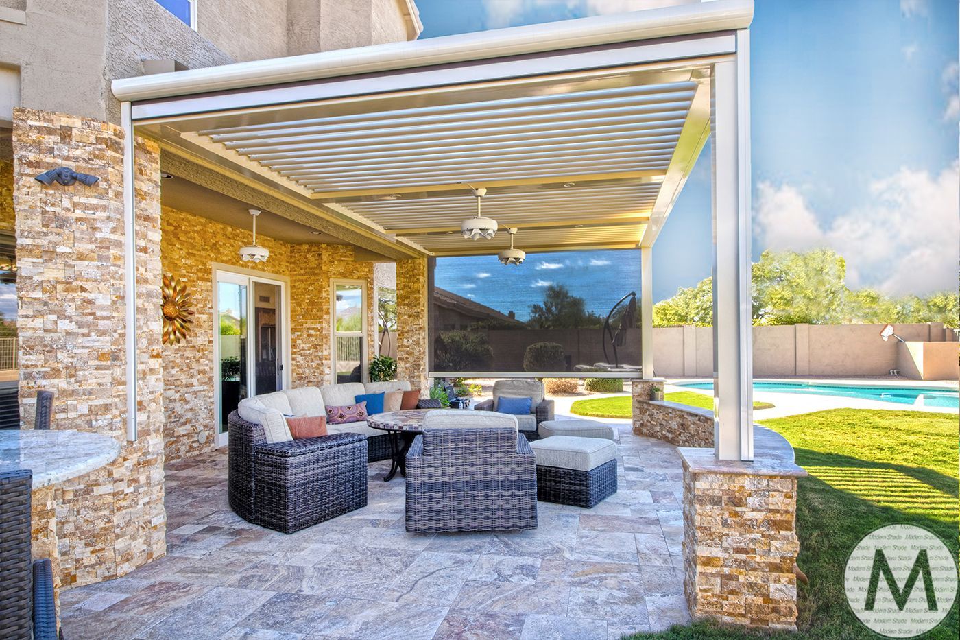 Patio with stone columns, wicker furniture, and a retractable roof overlooking a pool on a sunny day.