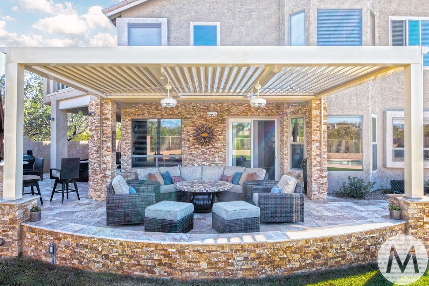 Patio with beige pergola, stone seating, and outdoor furniture in front of a two-story house.