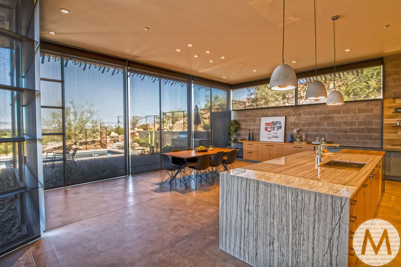 Modern kitchen with large windows, concrete floor, island, and pendant lights, overlooking desert landscape.