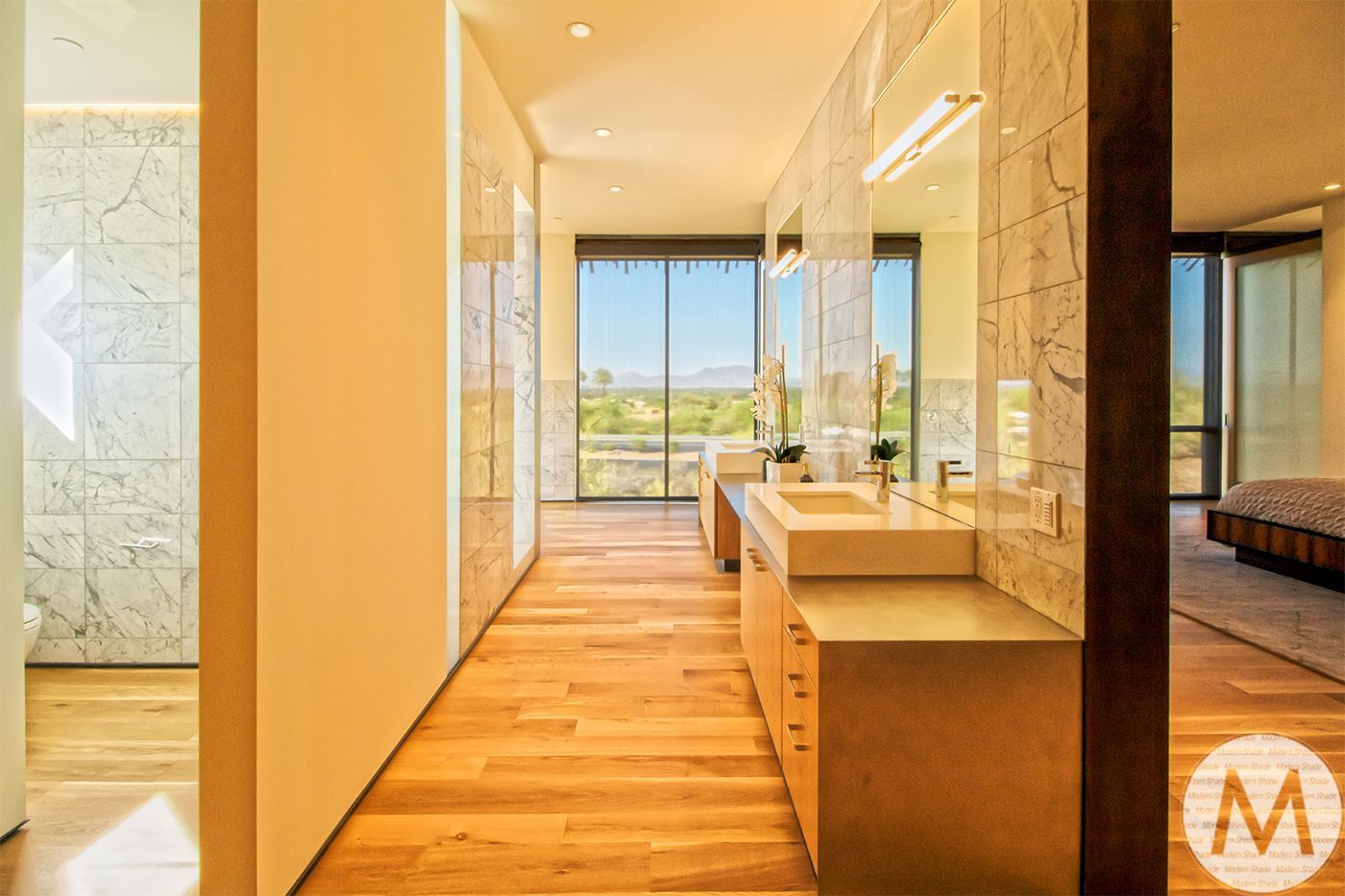 Bathroom with wood floor, marble walls, double sinks, and large window overlooking a scenic view.