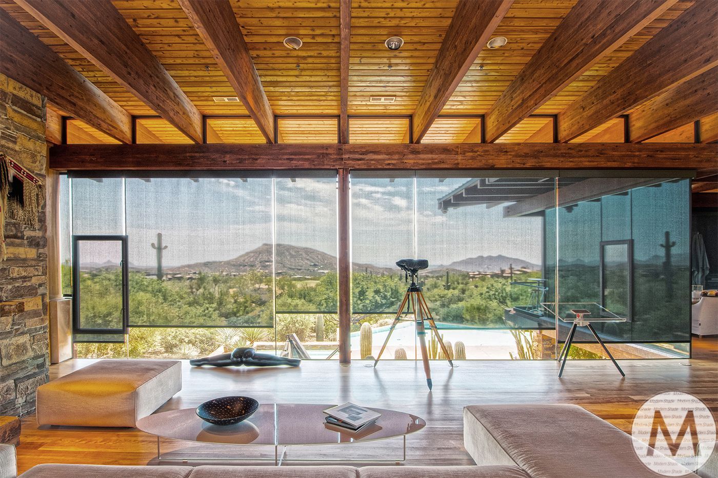 Living room with large windows overlooking a desert landscape and mountains; wooden ceiling and floor.