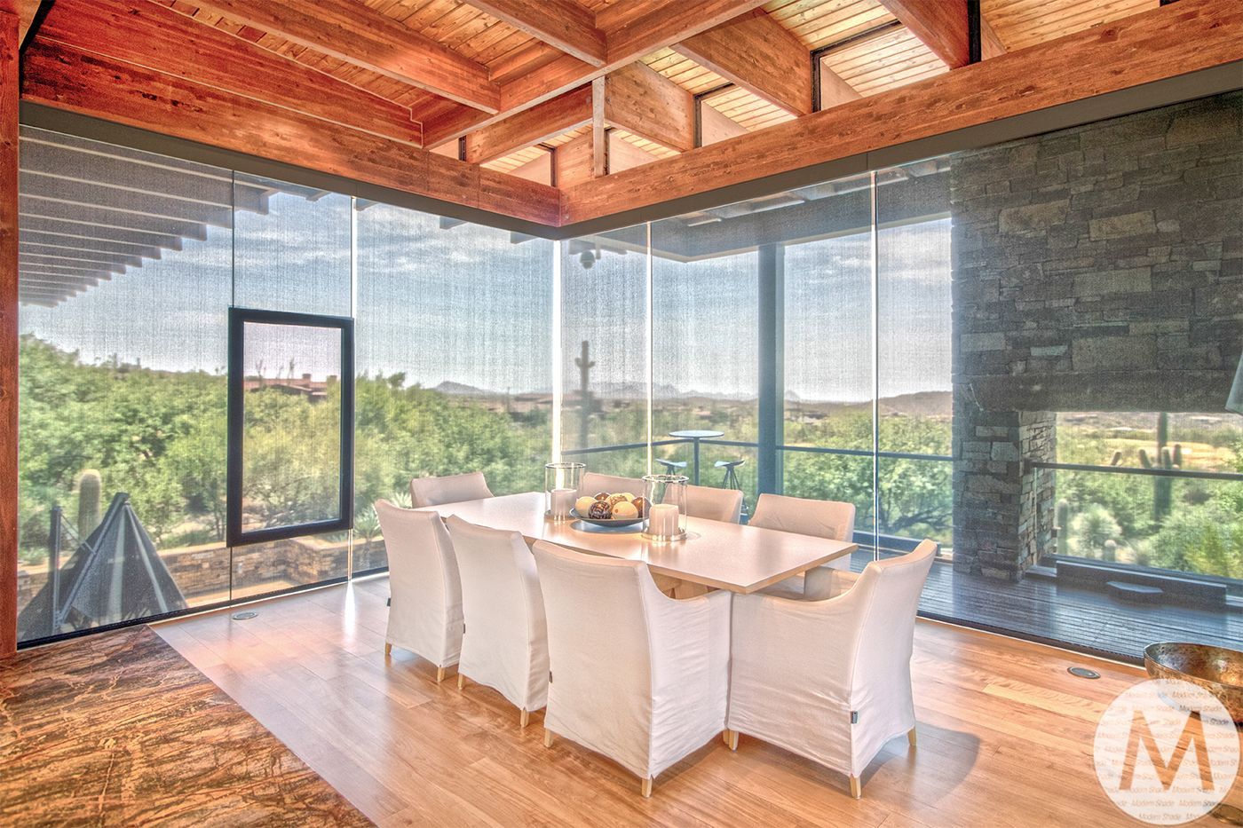 Dining room with large windows overlooking a landscape. Wooden beams, wood floor, table with white chairs.