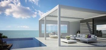 A white pergola overlooks a pool and ocean. Outdoor furniture, white walls, and a sunny sky.