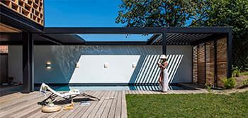 Poolside scene: A person in a white outfit stands by a pool under a pergola. Wooden deck, green lawn, and blue sky.