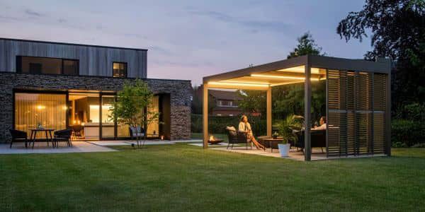 A modern house with illuminated backyard pergola and people gathered around seating.