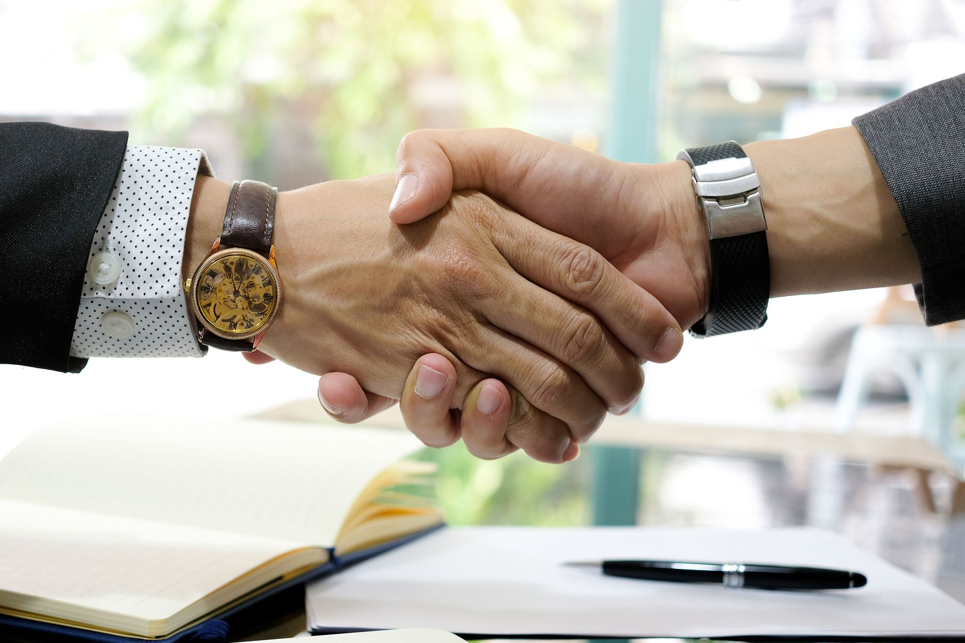 Two people in suits shake hands over a notepad and pen, in a business setting.