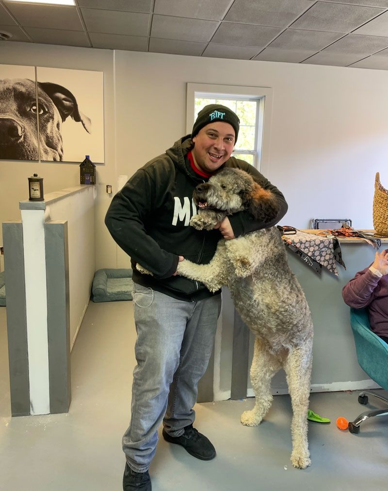 Man hugging a large, fluffy dog indoors. The man wears a black hoodie, hat, and gray pants. The dog is gray and white.