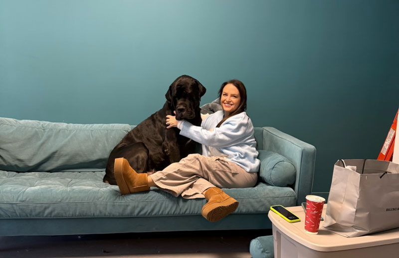 Woman in light blue sweatshirt cuddles a large black dog on a teal couch. Teal wall background.