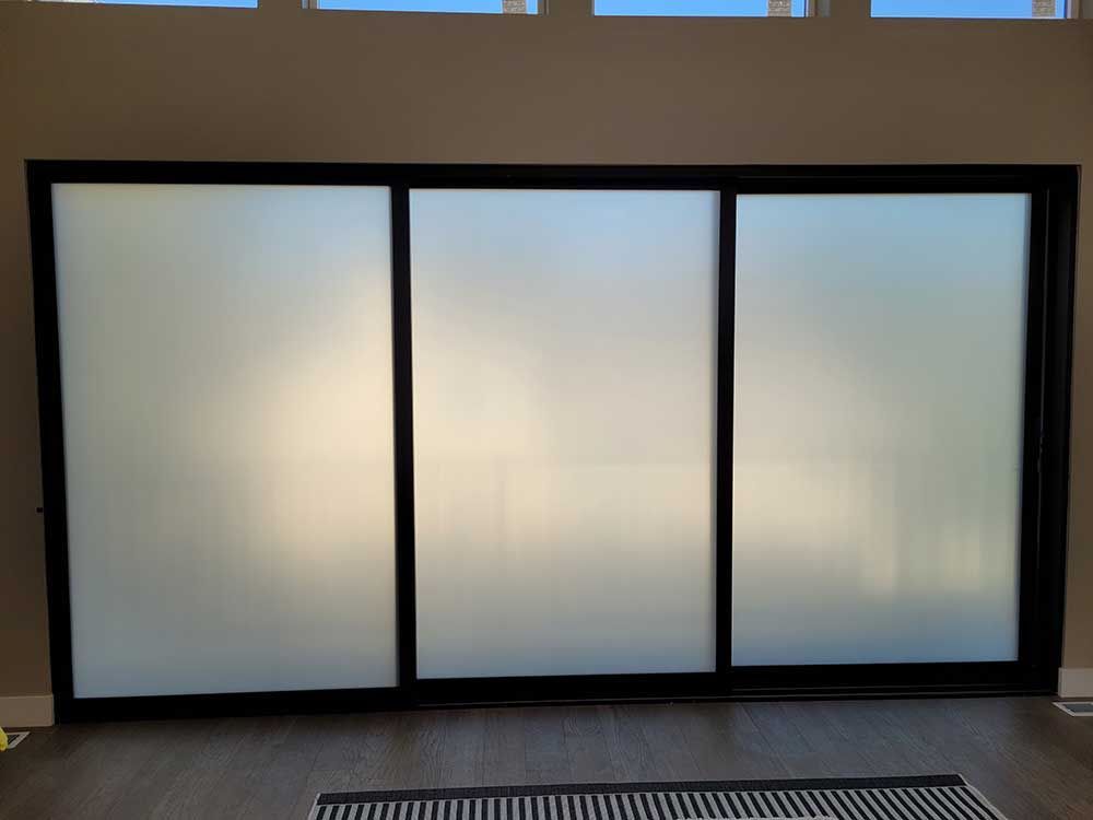 Three-panel sliding glass door with frosted glass and black frame. The door is set in a neutral-colored room.
