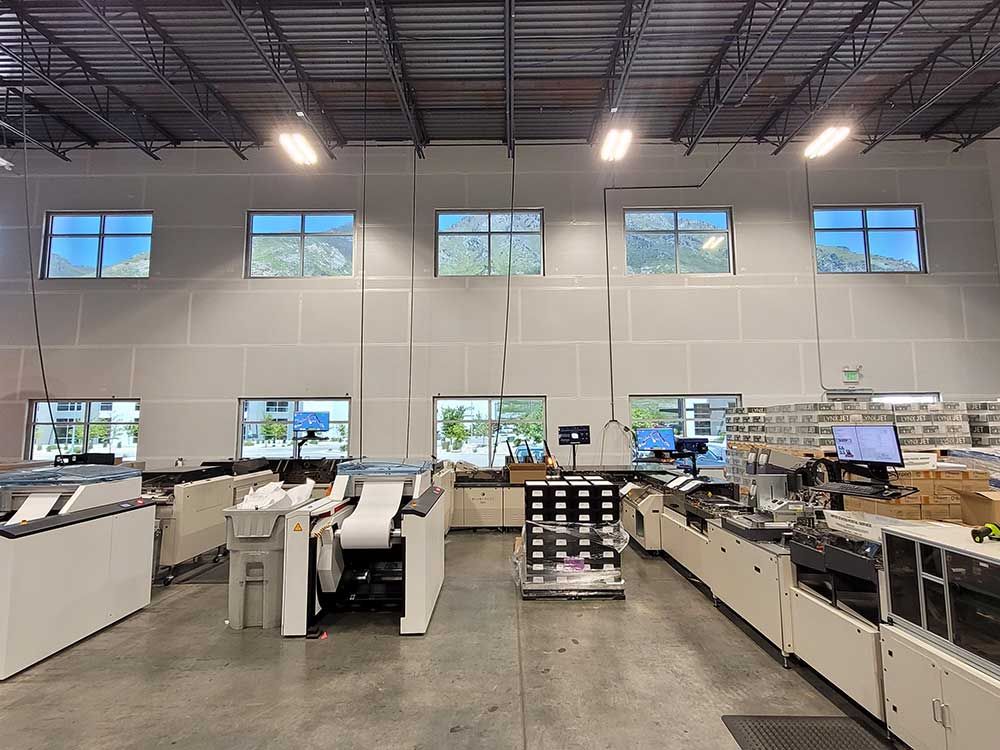 An industrial interior showing mail processing equipment. White machines are arranged in a row near a wall with windows and bright overhead lights.