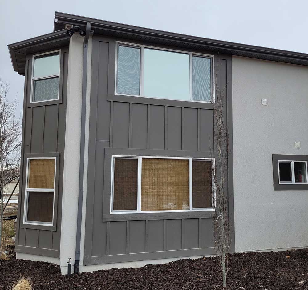 Two-story house exterior with gray siding and light gray stucco. Several windows with dark and light screens.